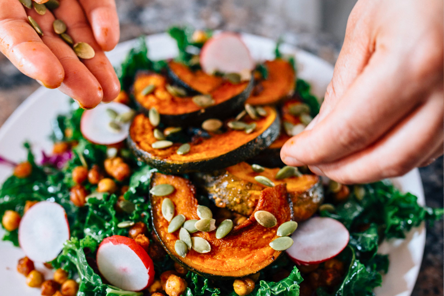 Hands adding roasted pumpkin seeds to a roasted squash salad.