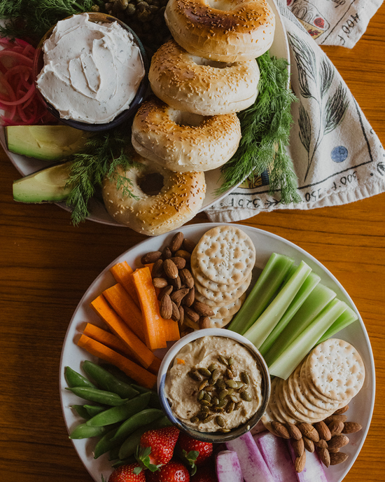 Brunch catering spread with bagels, vegan lox, crudites, crackers, nuts, and hummus.